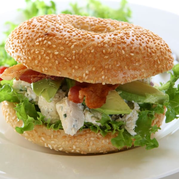 Bagel Nosh sesame bagel with chicken salad, bacon, avocado, and lettuce on a white plate.