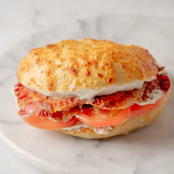A Bagel Nosh bagel with melted cheese, bacon, cream cheese, and sliced tomatoes on a marble surface.