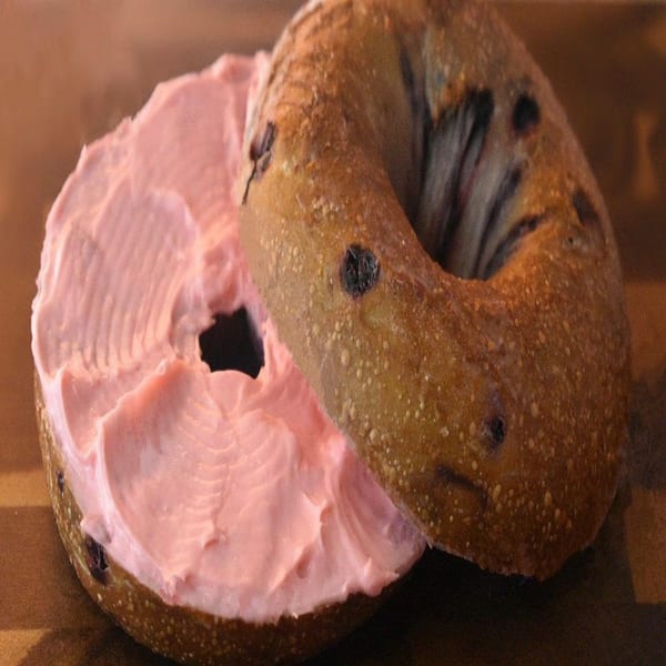 A blueberry bagel from Bagel Nosh, sliced in half with pink cream cheese spread on one side.