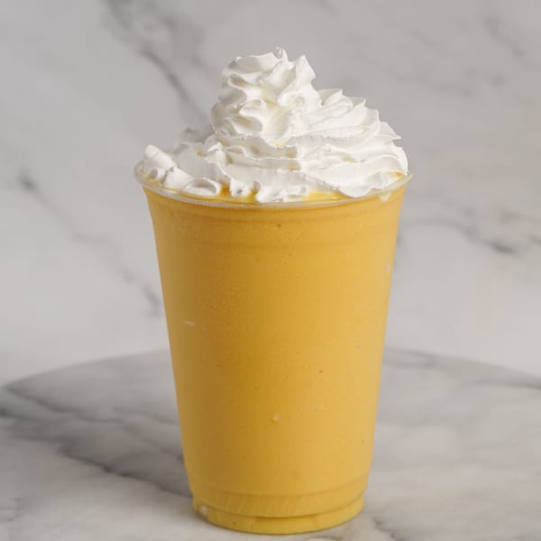 A yellow smoothie topped with a generous swirl of white whipped cream in a clear plastic cup.