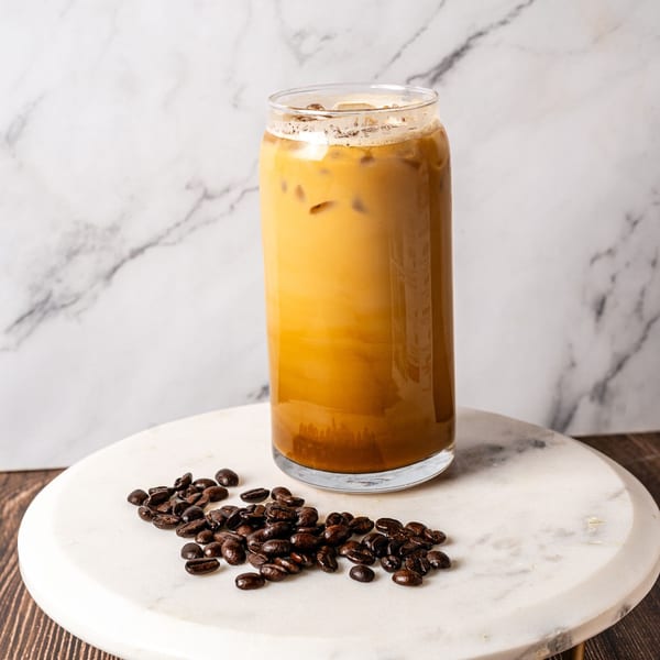 Iced coffee in a tall glass with coffee beans scattered on a marble surface.