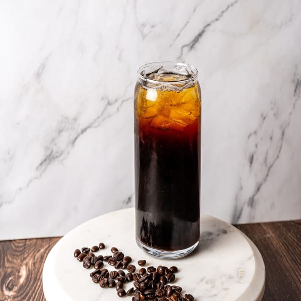 Iced coffee in a tall glass with coffee beans scattered on a marble surface.