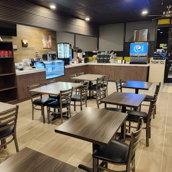 Interior of Bagel Nosh with empty tables and chairs, a counter with a display case, and a Pepsi machine.
