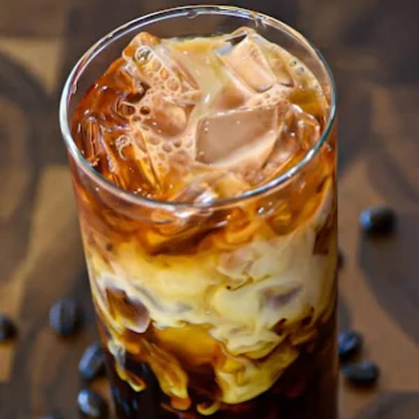 Iced coffee with cream and ice in a tall glass, with coffee beans scattered around.