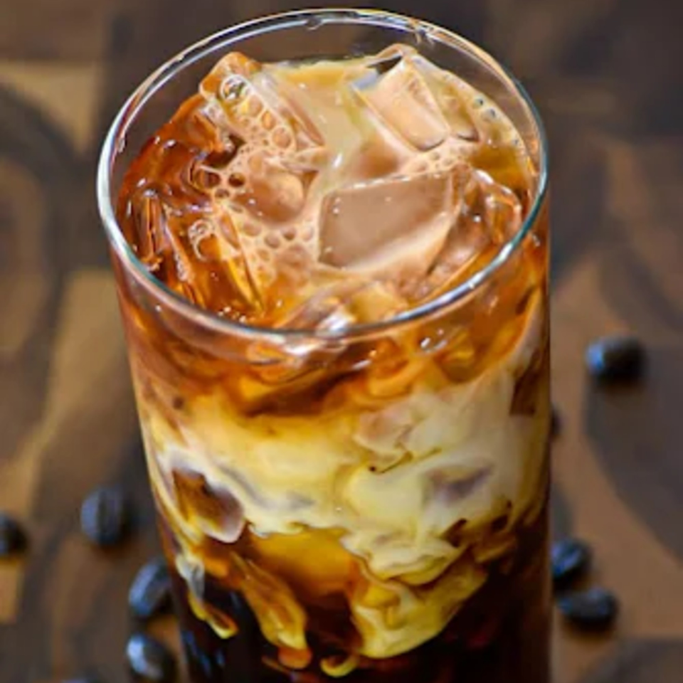Iced coffee with cream and ice in a tall glass, with coffee beans scattered around.