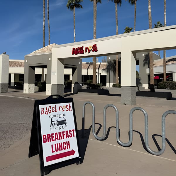 Bagel Nosh storefront with a curbside pickup sign, bike rack, and palm trees under a blue sky.