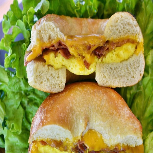 Two Bagel Nosh breakfast bagels, cut in half, filled with egg, bacon, and melted cheese on lettuce.