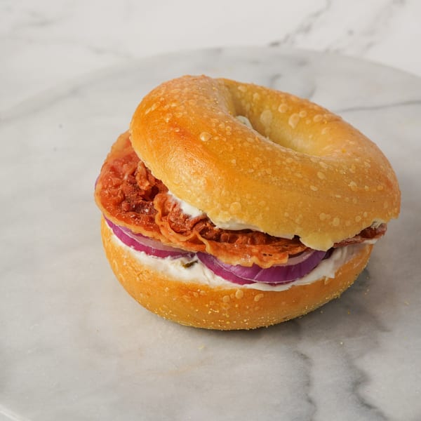 Bagel Nosh bagel with cream cheese, crispy bacon, and red onion slices on a marble surface.